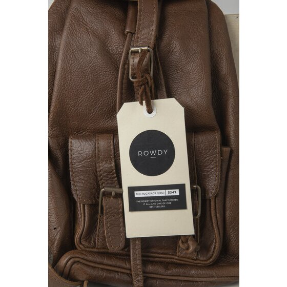 Large buff tag printed with a round ROWDY logo hanging from the buckle strap of a brown leather backpack