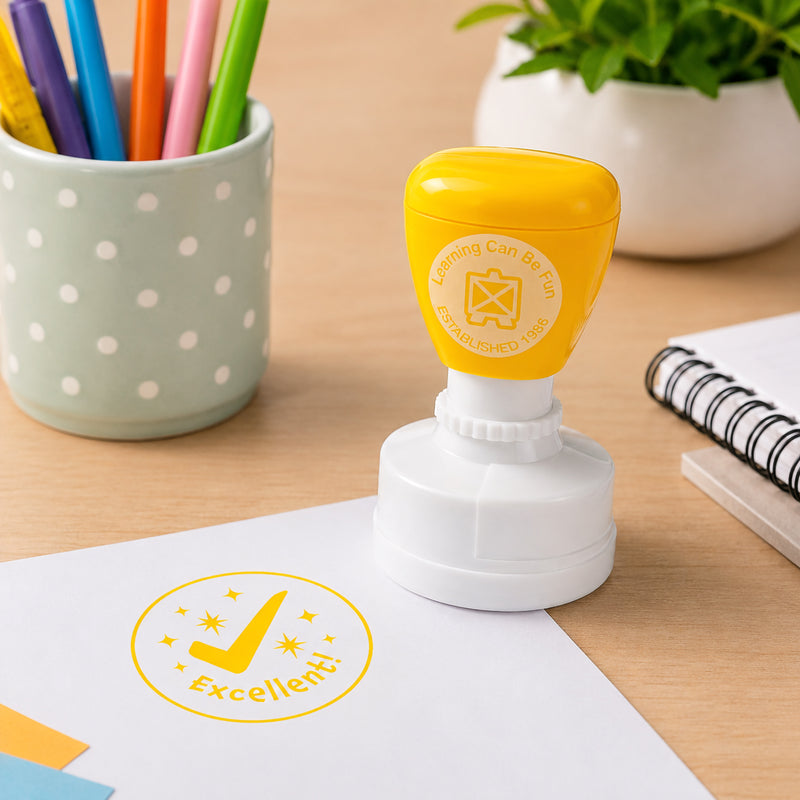 Excellent merit stamp positioned next to stationery and classroom supplies with a crisp yellow tick design stamped onto paper. The encouraging message provides positive reinforcement for student achievement and effort.