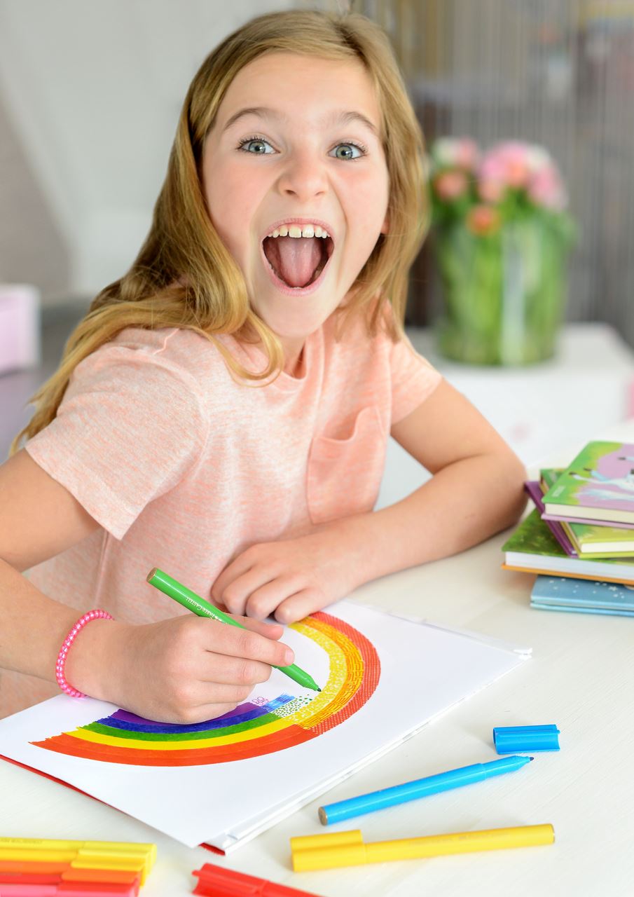 Smiling child colouring a rainbow with Faber-Castell Connector felt tip pens, with more pens and caps spread across the table