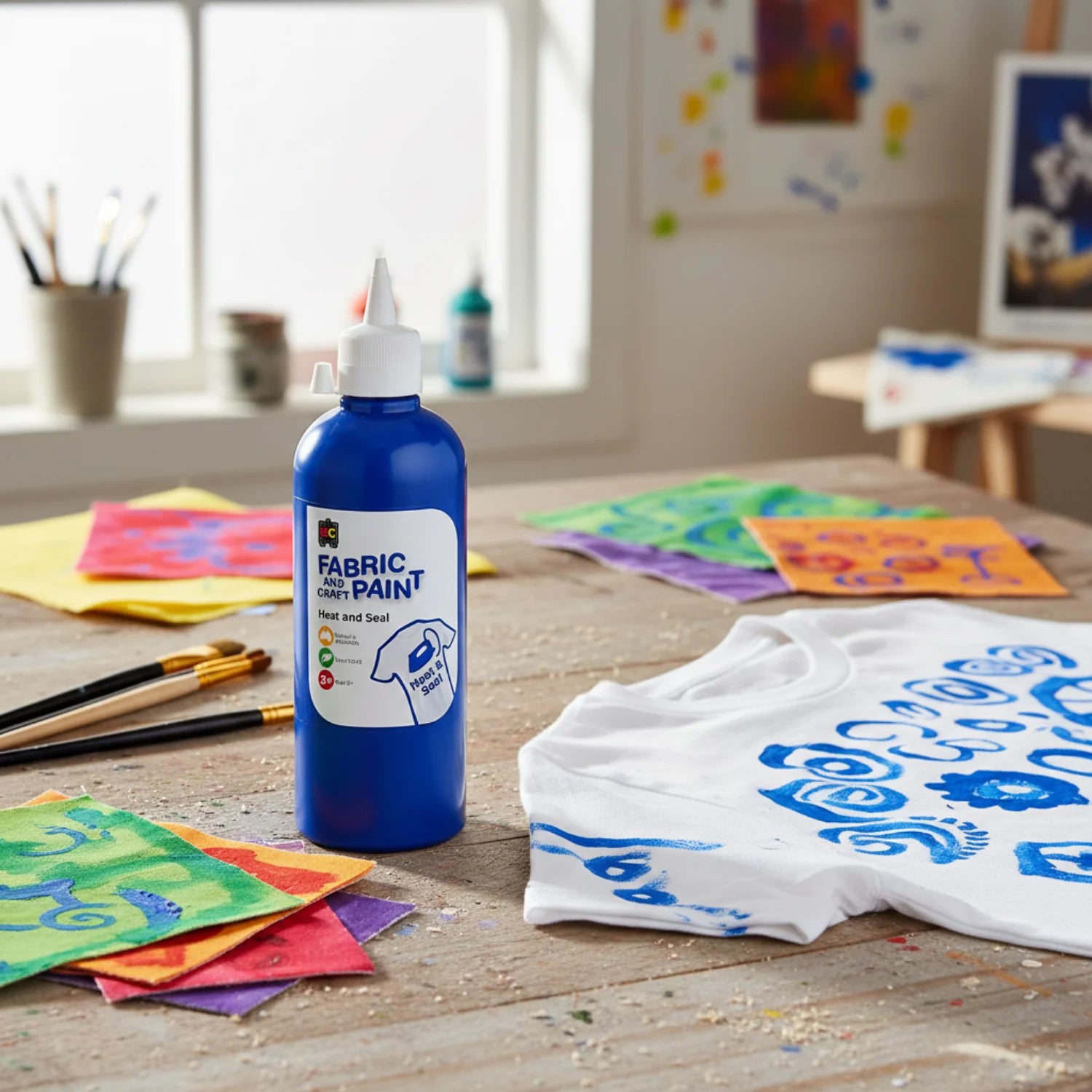 Educational Colours Fabric and Craft Paint 500ml Dark Blue displayed on a rustic art table surrounded by colourful painted fabric swatches, paint brushes, and a white shirt with blue decorative artwork.