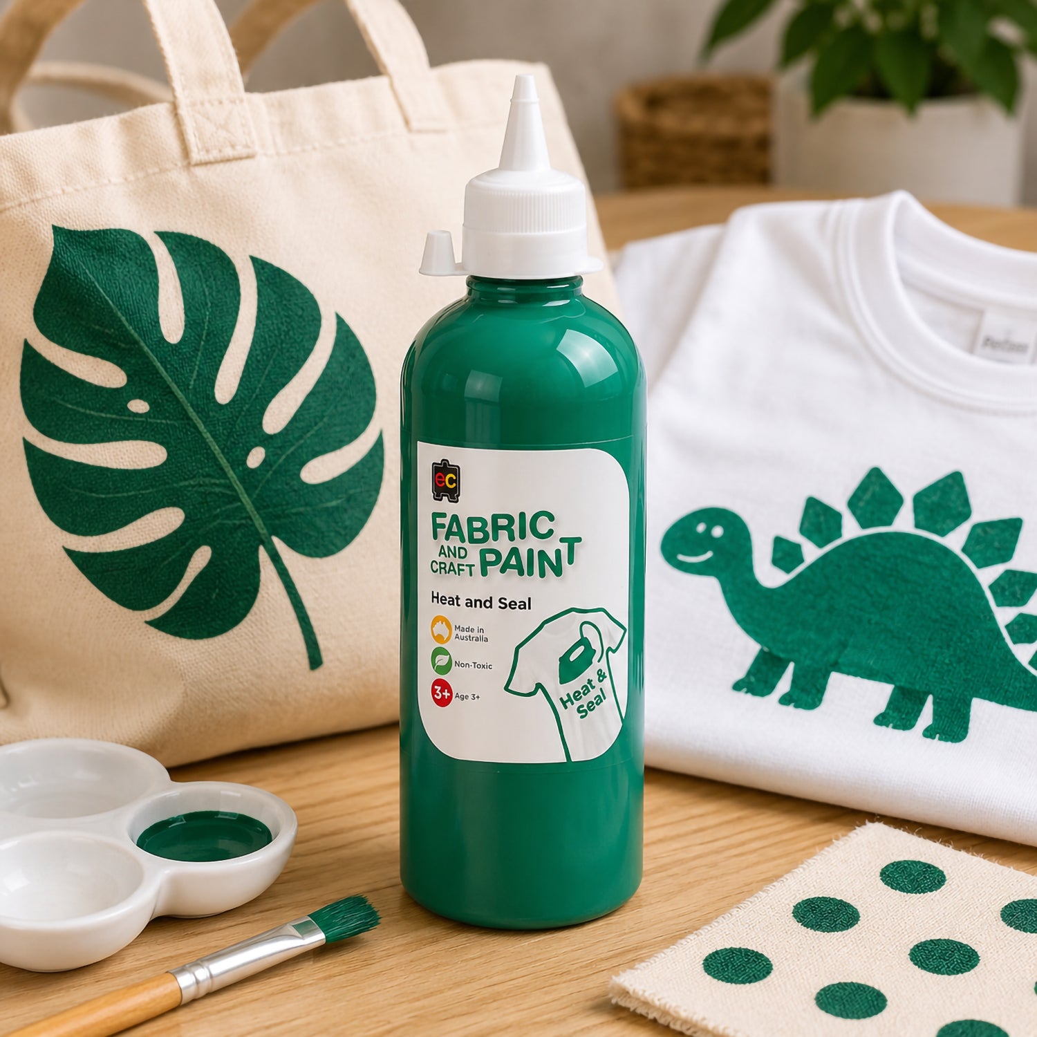 Educational Colours forest green fabric paint shown beside a tote bag with tropical leaf artwork and a white shirt decorated with dinosaur themed green designs.