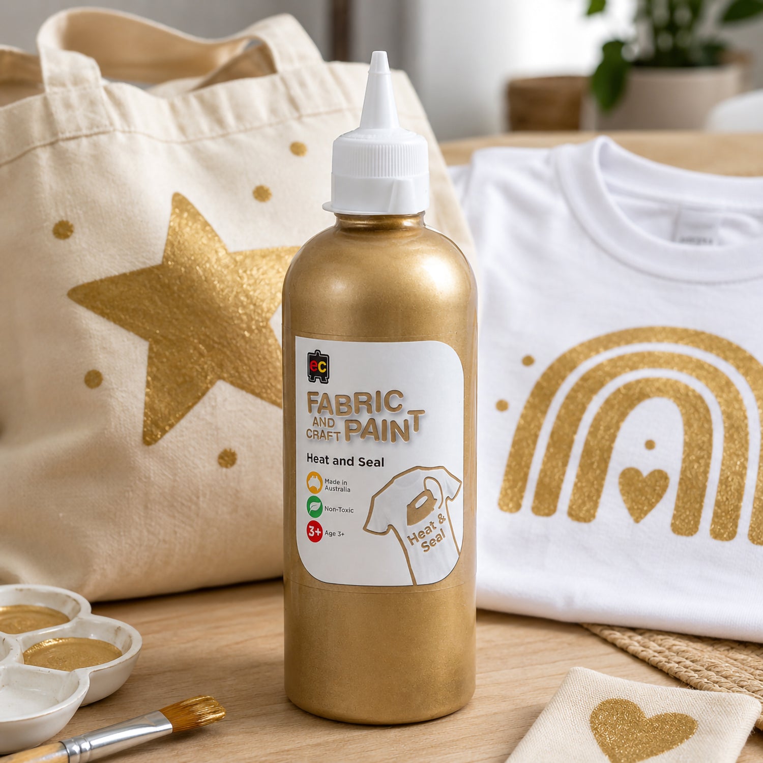 Educational Colours gold fabric paint shown beside a decorated tote bag and white shirt featuring bright metallic gold painted shapes and patterns in a creative studio setting.