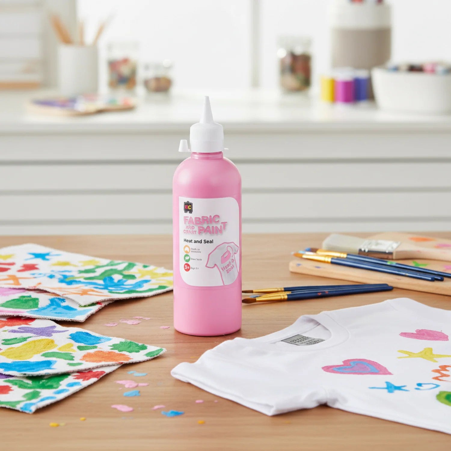 Educational Colours pink craft paint bottle standing on a craft table with colourful painted fabric pieces, paint brushes, and decorated clothing projects in a bright creative workspace.