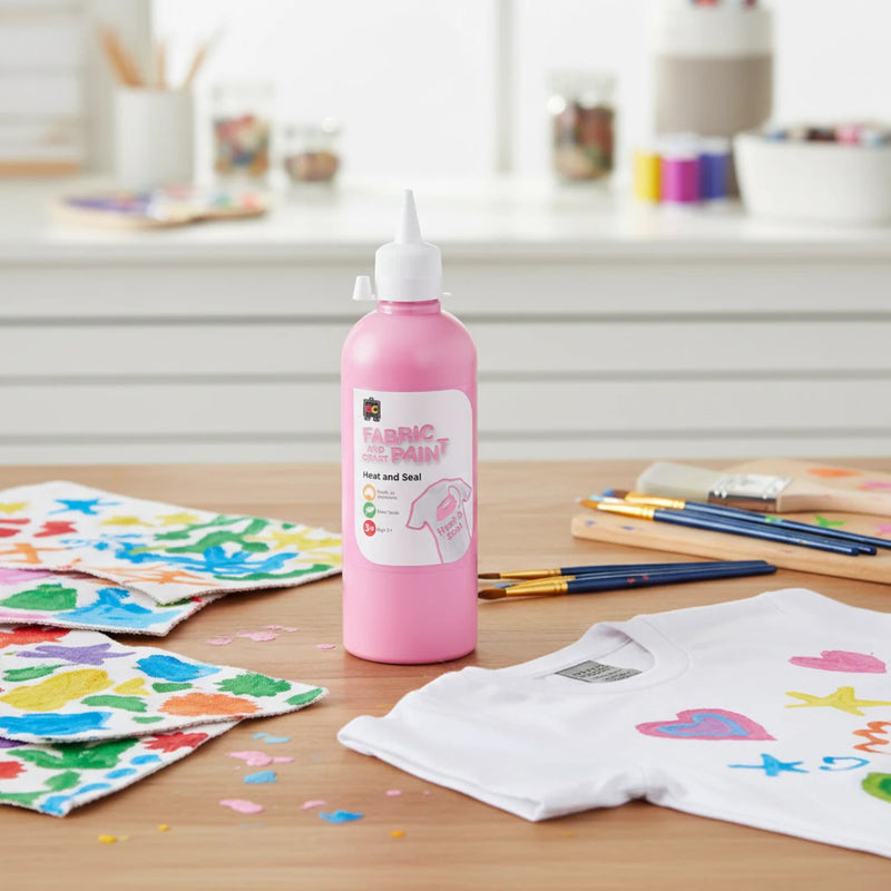 Educational Colours pink craft paint bottle standing on a craft table with colourful painted fabric pieces, paint brushes, and decorated clothing projects in a bright creative workspace.