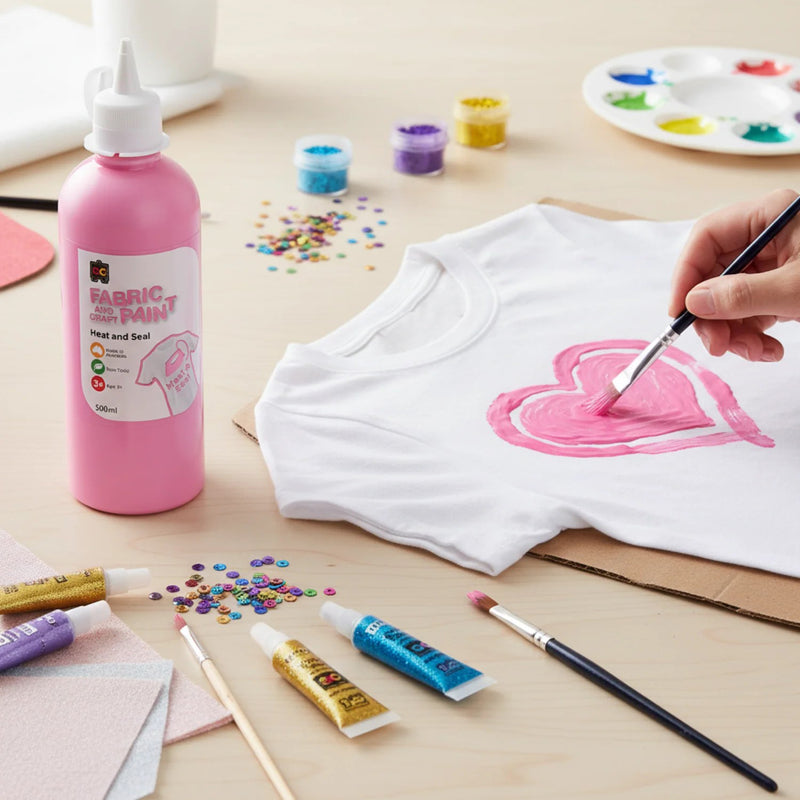 Pink fabric and craft paint bottle positioned next to a hand painting a pink heart design on a white t shirt surrounded by sequins, brushes, and colourful craft supplies.