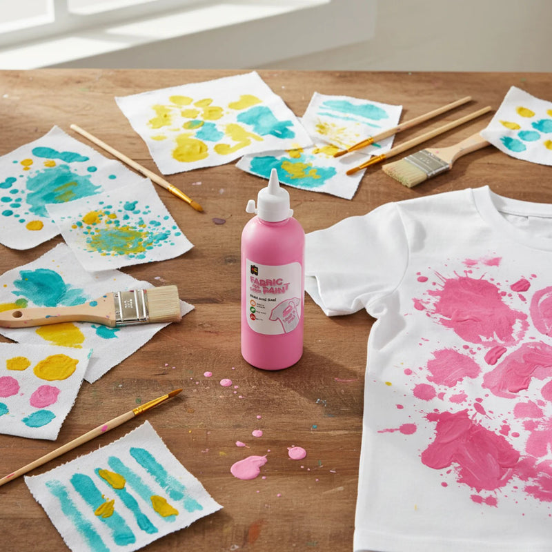 Educational Colours Fabric and Craft Paint 500ml Pink displayed on a wooden craft table beside painted fabric samples, brushes, and a white t shirt featuring bright pink painted designs.