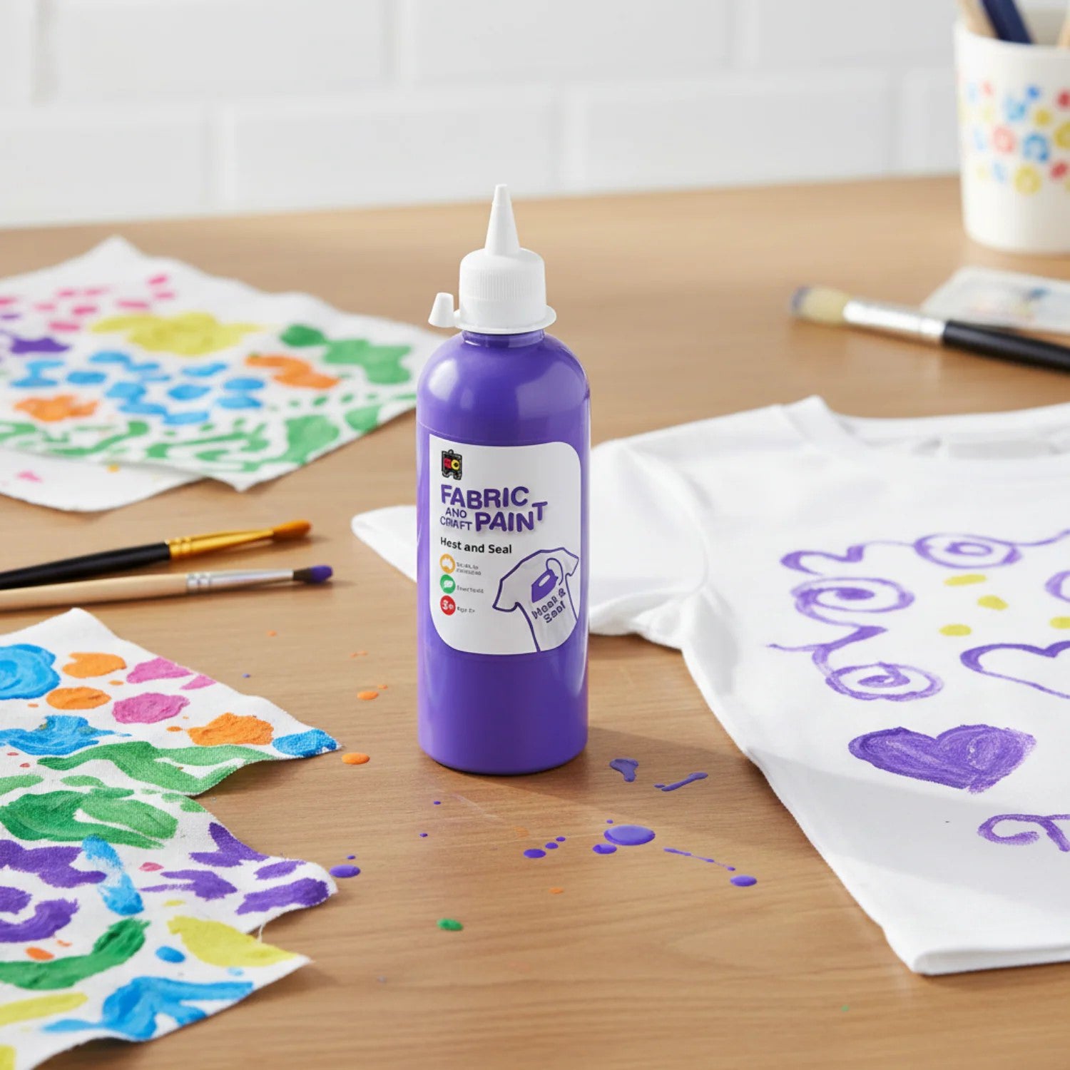 Bottle of purple fabric and craft paint positioned with colourful painted cloth pieces and a decorated white shirt featuring purple painted patterns and hearts for fabric art inspiration.