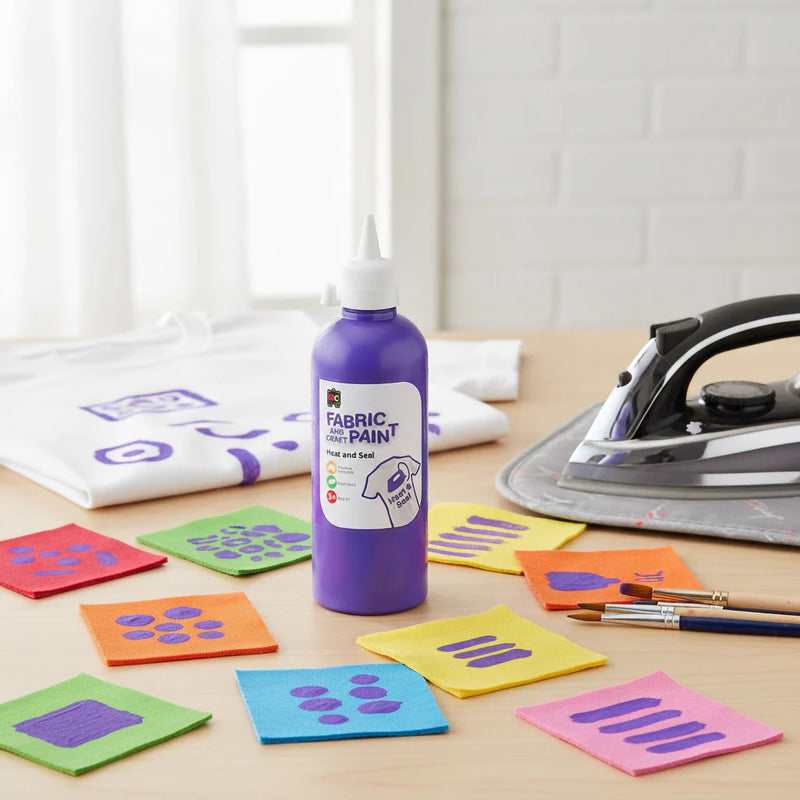 Purple Educational Colours Fabric and Craft Paint bottle beside painted fabric swatches, brushes, and an iron used for heat sealing textile artwork in a bright classroom craft environment.