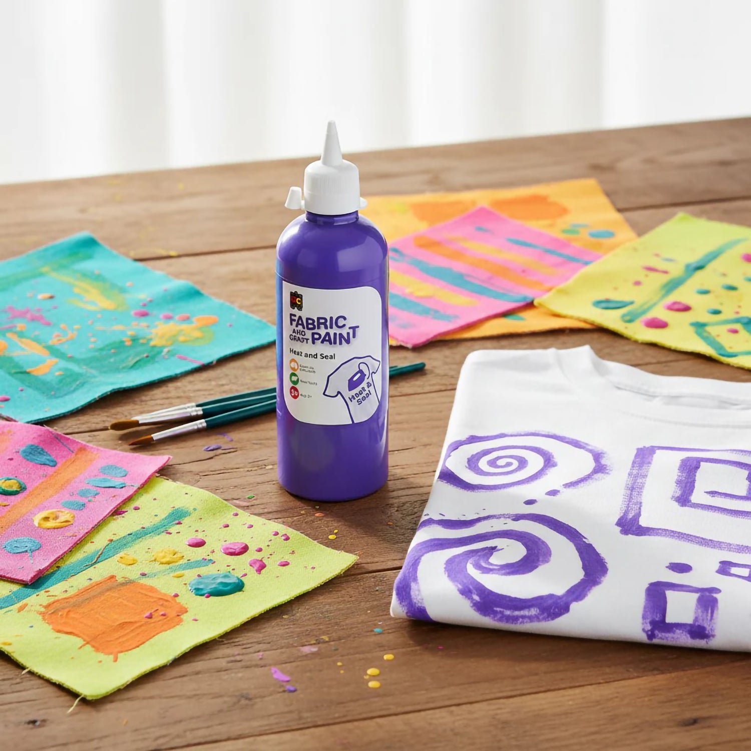 Educational Colours Fabric and Craft Paint 500ml Purple displayed on a wooden craft table surrounded by colourful painted fabric samples, paint brushes, and decorated textile projects showing creative fabric art applications.