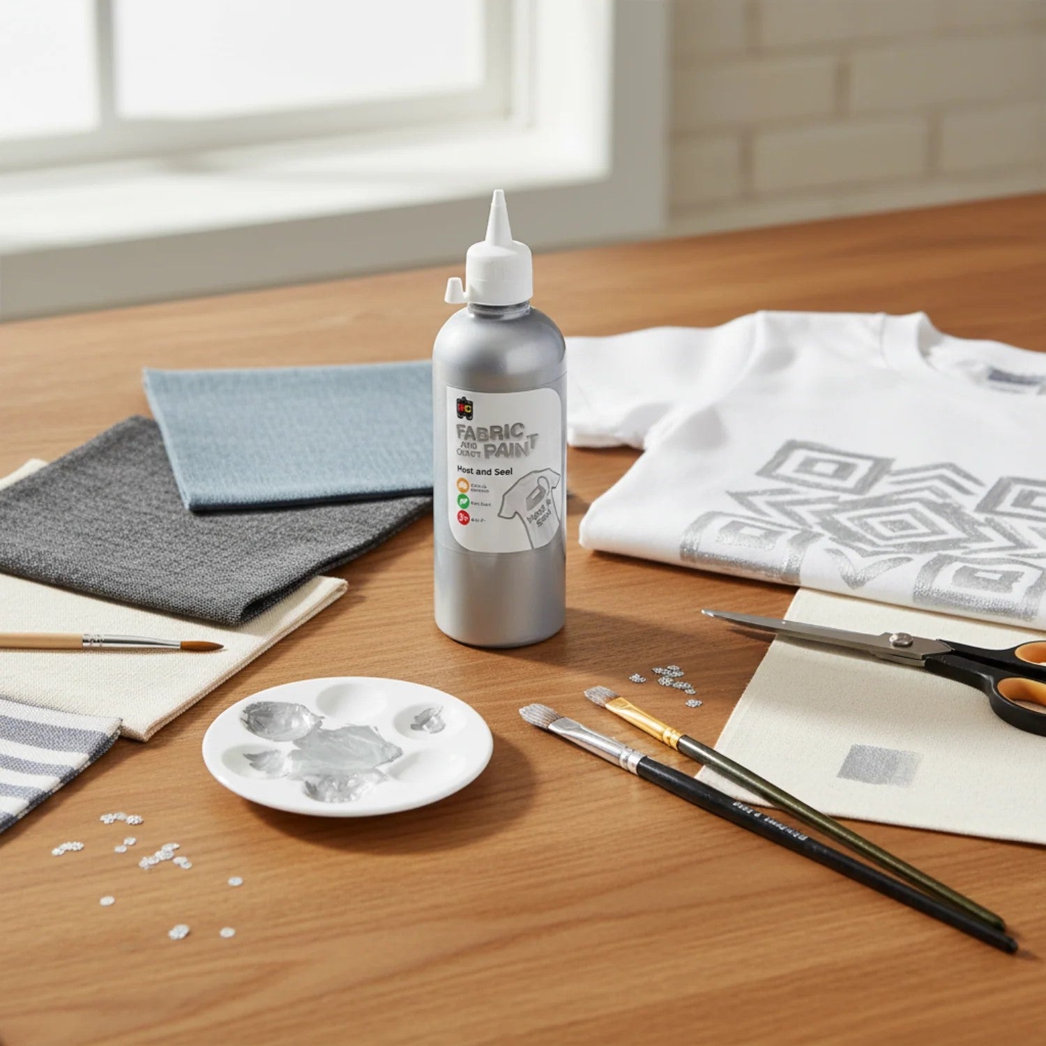 Educational Colours silver paint bottle shown in a creative workspace with fabric materials, paint brushes, sequins, and a white shirt decorated with geometric metallic silver patterns.