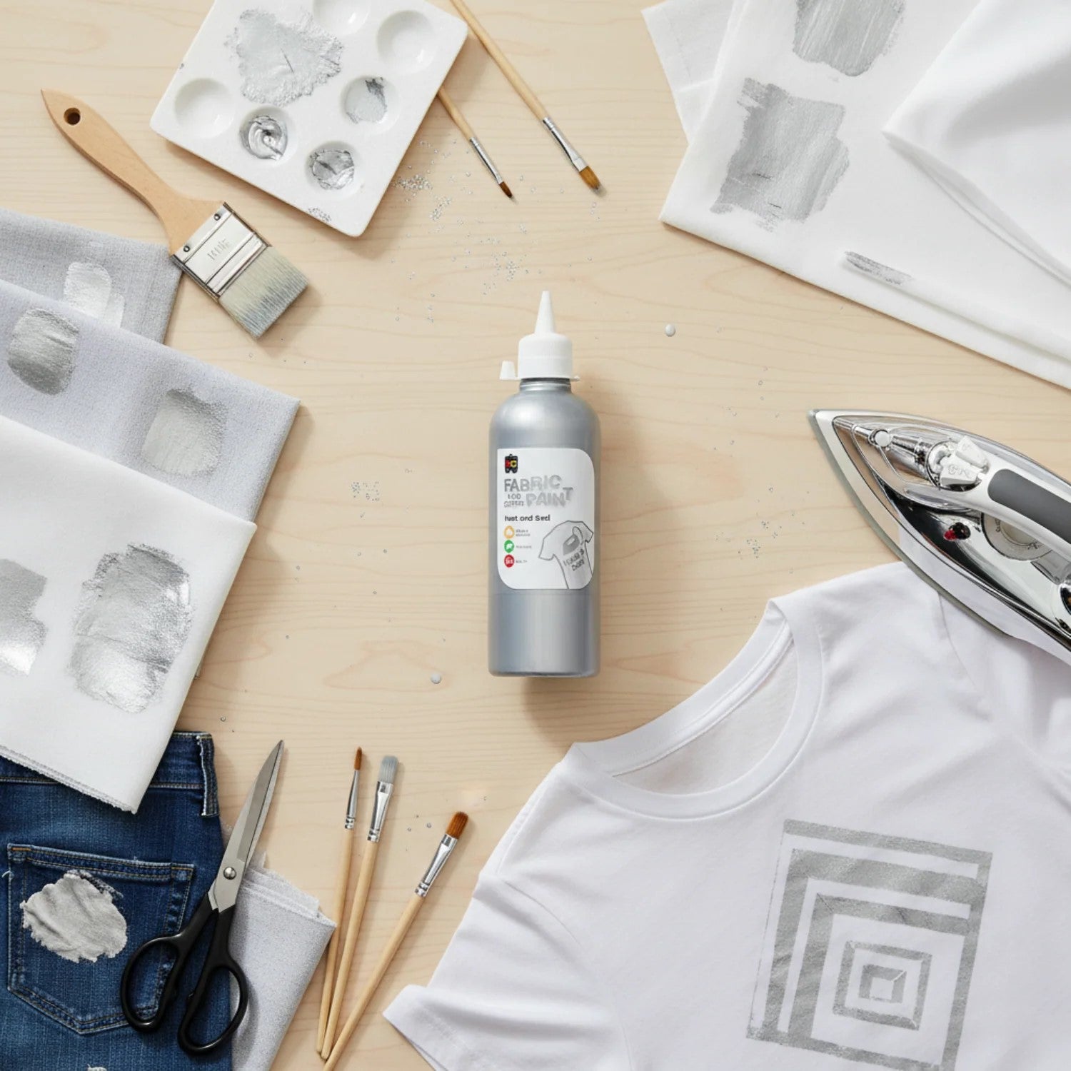 Silver fabric and craft paint bottle placed beside painted textile swatches, a paint palette, brushes, scissors, and an iron used for heat sealing fabric artwork.