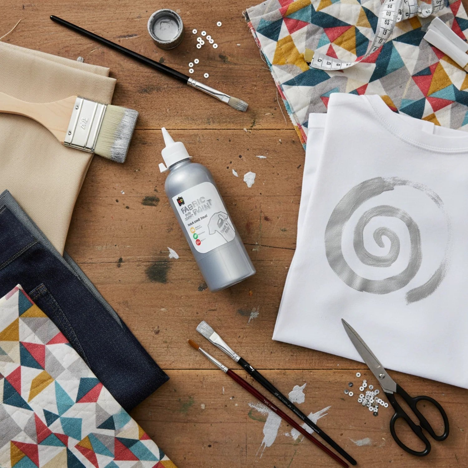 Educational Colours Fabric and Craft Paint 500ml Silver displayed on a wooden craft table with fabric samples, paint brushes, scissors, and a white t shirt featuring a metallic silver spiral design.