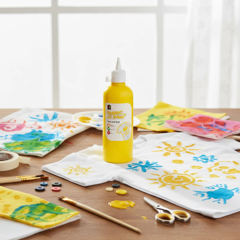 Yellow fabric and craft paint bottle positioned beside decorated t shirts, painted fabric pieces, brushes, scissors, and vibrant mixed colour craft projects on a wooden workspace.