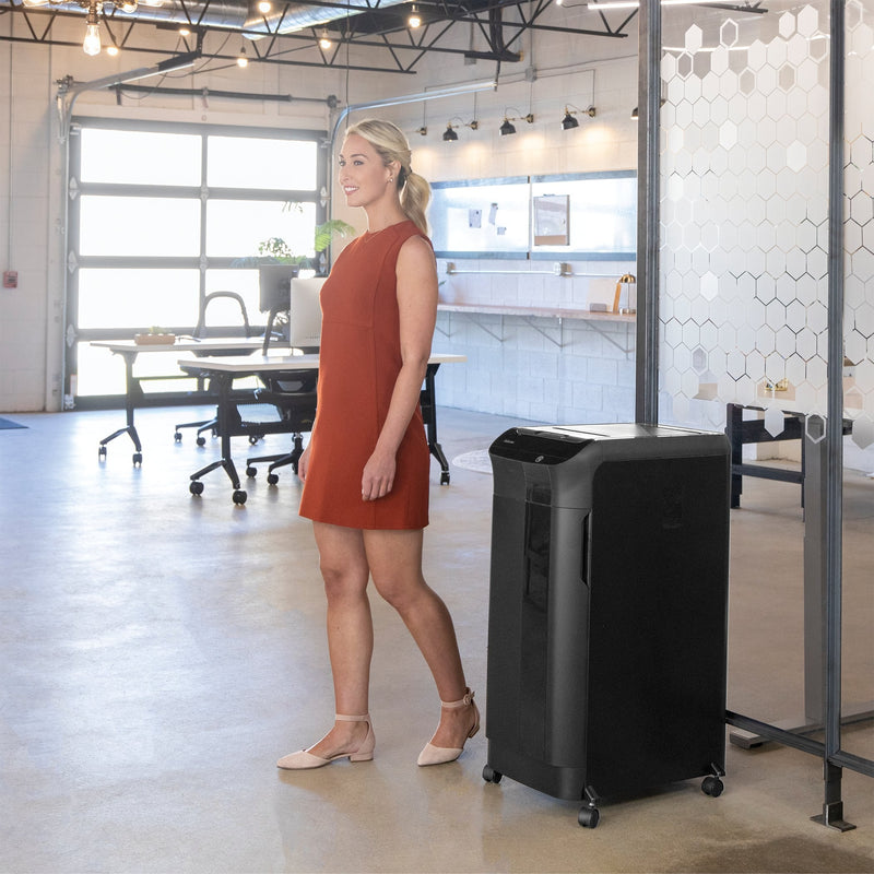 Woman in a red dress walking past a tall black Fellowes AutoMax cabinet shredder in a bright open-plan office