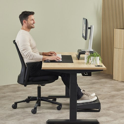 Man at a sit-stand desk typing with the black Breyta wrist rest in front of his keyboard, a footrest under the desk