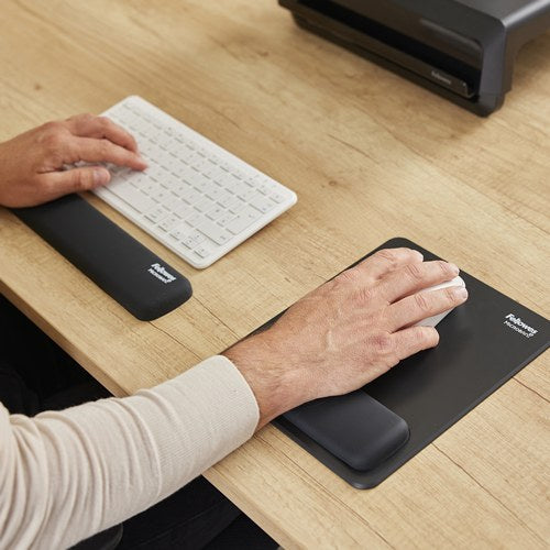 Hand on a white mouse resting on a black Breyta mouse pad wrist rest beside a black Breyta keyboard rest on a timber desk