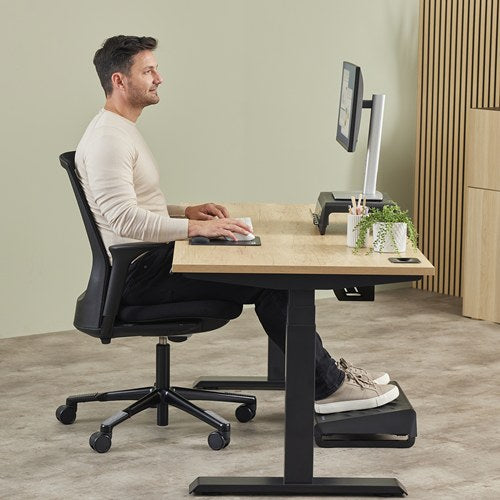 Man seated at a timber sit-stand desk using a mouse on a black Breyta pad, with a footrest under the desk