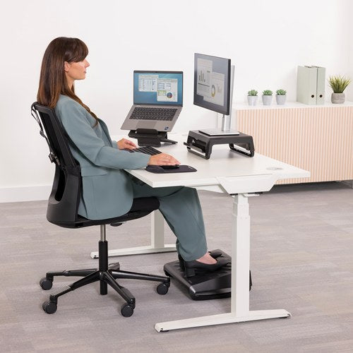 Woman working at a white sit stand desk with laptop and monitor, her feet on the Breyta foot rest