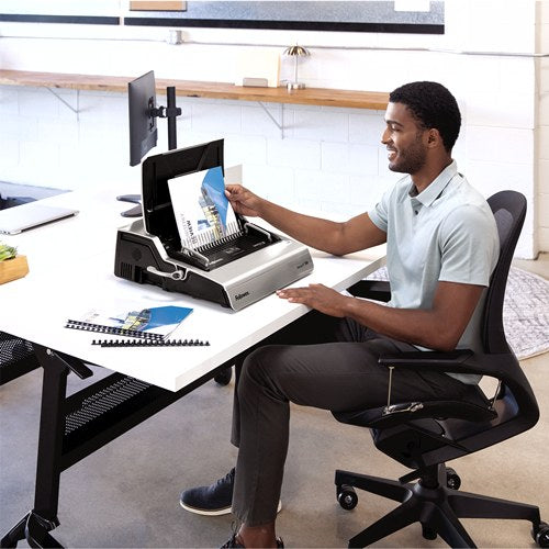Man at a desk feeding a document into a Galaxy-E comb binder, bound reports and combs spread beside him