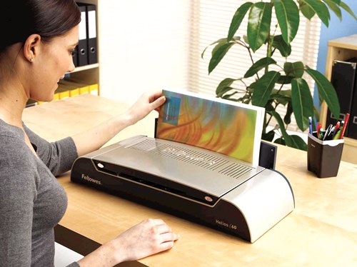 Woman at an office desk standing a bound document in the open Helios 60 thermal binder, shelves and a pot plant behind