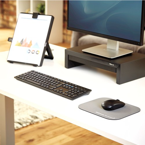 Graphite Fellowes pad and black mouse on a white desk beside a keyboard and black monitor riser