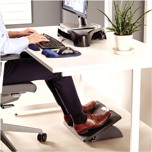 Man in a shirt typing at a white desk with both feet in brown shoes resting on the black Office Suites footrest