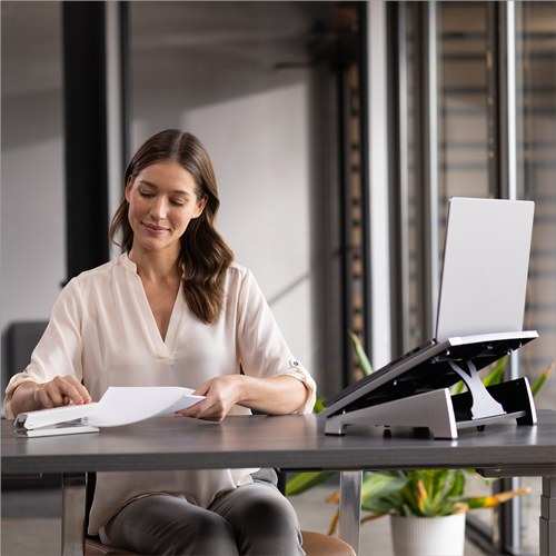 Woman reading papers at a desk with her laptop raised on a silver and black Office Suites laptop riser