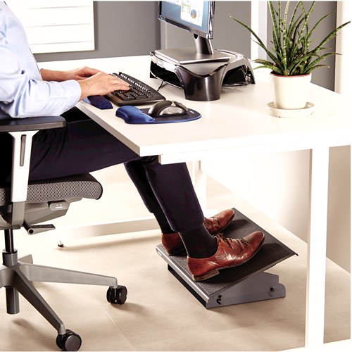 Man seated at an office desk with both feet in brown shoes resting on the steel heavy duty foot support