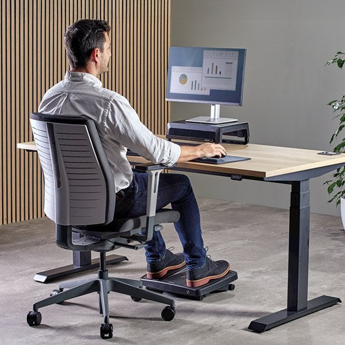 Man working at a sit stand desk with a monitor, seated with his feet resting on the Ultimate foot support