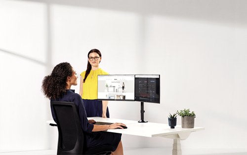 Two women at a white desk, one seated in front of two screens on a Fellowes Reflex dual arm