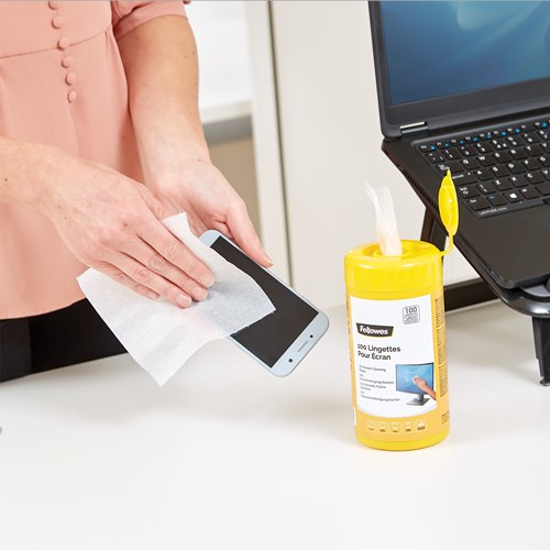 Hands wiping a smartphone screen with a Fellowes wipe, the yellow tub and a laptop on the desk behind