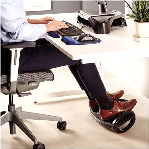 Man seated at a desk with keyboard and mouse, both feet in brown shoes resting on the black foot rocker