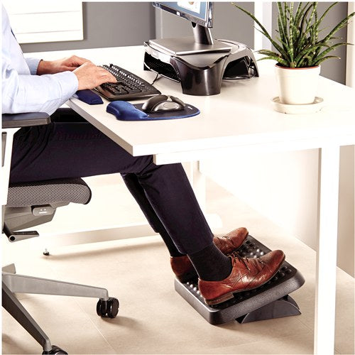 Man typing at a white desk with both feet in brown shoes on the graphite Standard Footrest