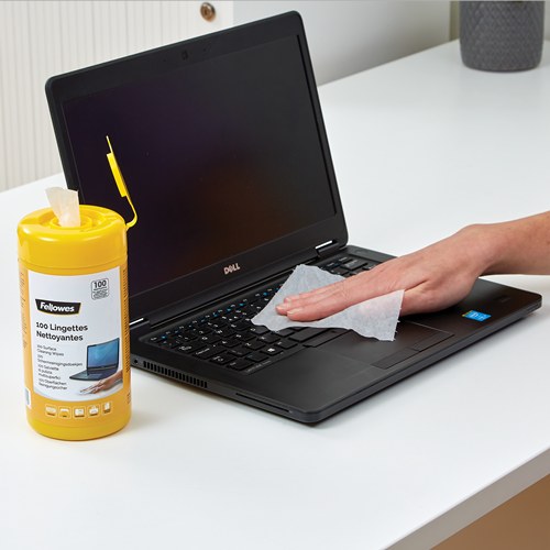 Hand wiping a black laptop keyboard with a white wipe, a yellow Fellowes surface wipes tub standing beside the laptop