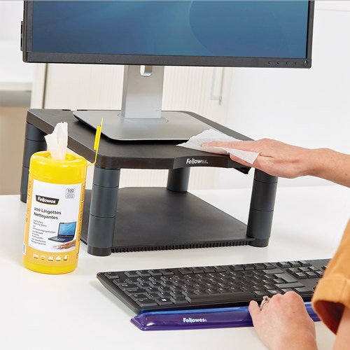 Hand wiping the top of a black monitor riser with a Fellowes wipe, the yellow tub and a keyboard on the desk