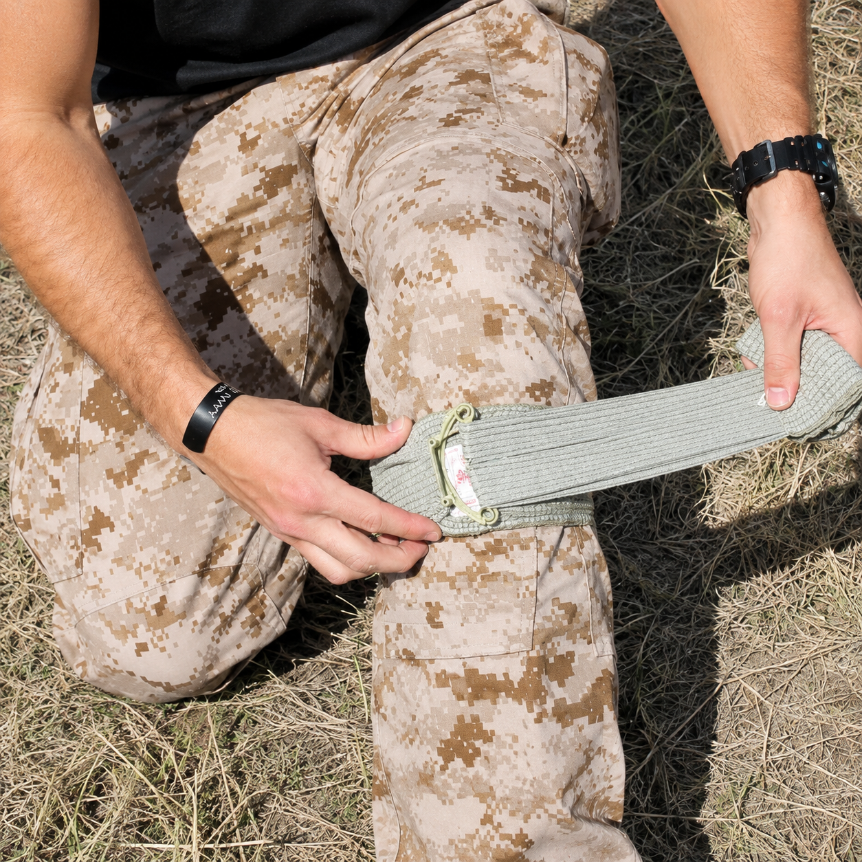 Person applying the FirstCare Military Trauma and Haemorrhage Control Bandage 15 x 18cm Green around the leg in an outdoor field setting. The image shows the elastic wrap being pulled across the dressing and tensioned around the limb, demonstrating practical application where rapid bleeding control and secure compression are required.