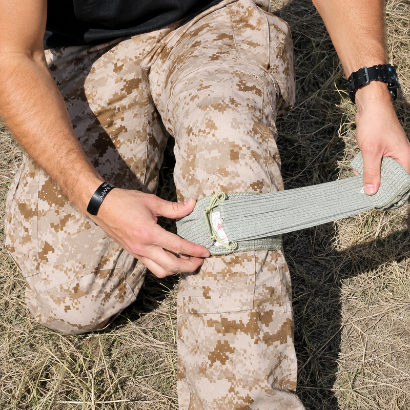 Person applying the FirstCare Military Trauma and Haemorrhage Control Bandage 15 x 18cm Green around the leg in an outdoor field setting. The image shows the elastic wrap being pulled across the dressing and tensioned around the limb, demonstrating practical application where rapid bleeding control and secure compression are required.