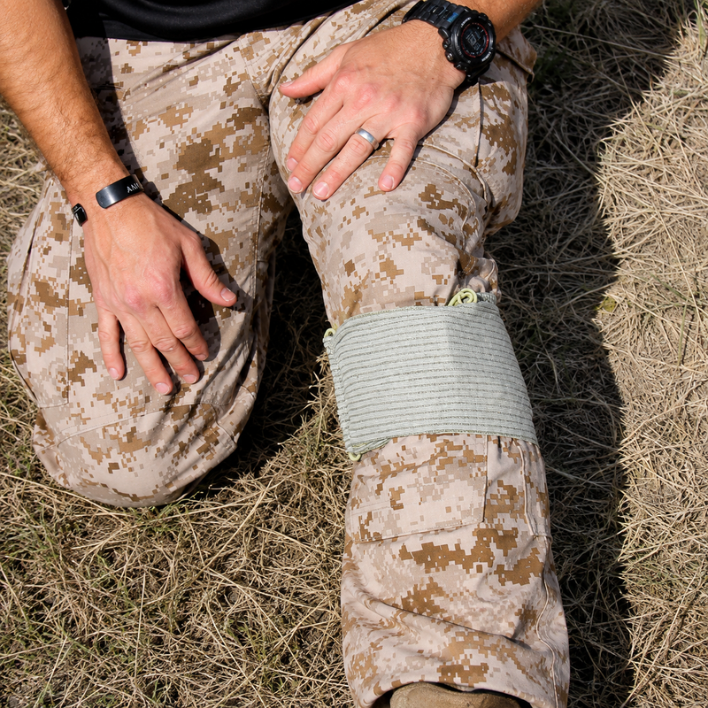 FirstCare Military Trauma and Haemorrhage Control Bandage 15 x 18cm Green secured around the upper lower leg of a person wearing field clothing. The lifestyle scene demonstrates the finished bandage configuration with the wide elastic wrap providing firm circumferential coverage around the injured area during an outdoor emergency response scenario.