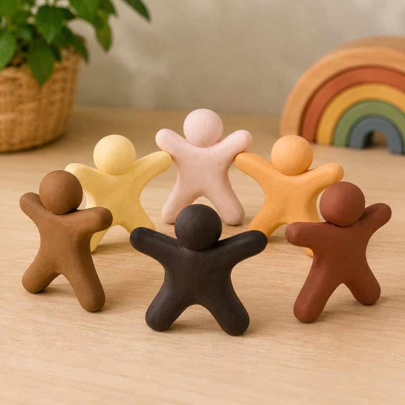 Six clay figures in different skin tone colours arranged in a circle on a wooden tabletop. The figures represent diversity, inclusion and friendship, showcasing the six colours included in the Friendship Colours Creative Clay set against a warm educational setting.