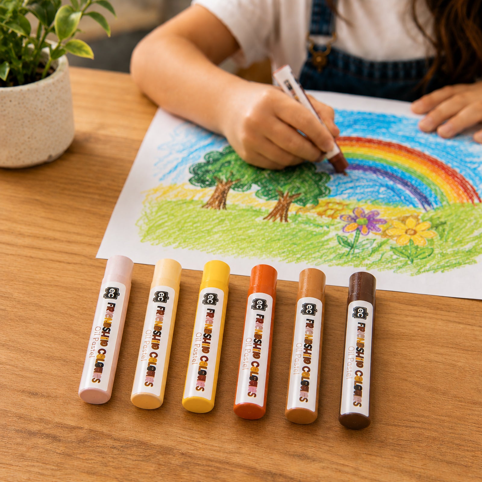 Child using Friendship Colours Oil Pastels to colour a vibrant rainbow landscape featuring trees, flowers and a bright sky. Six pastel shades are displayed in the foreground, demonstrating how the smooth oil pastels encourage creative expression, colour exploration and artistic confidence in educational settings.