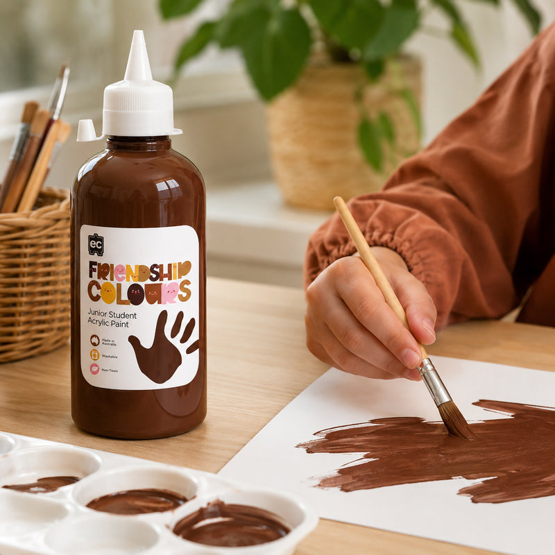 Bottle of Friendship Colours Espresso Acrylic Paint positioned next to paint palettes filled with deep brown paint. A student paints broad brushstrokes on paper while classroom art tools and natural surroundings create a warm and creative learning environment.