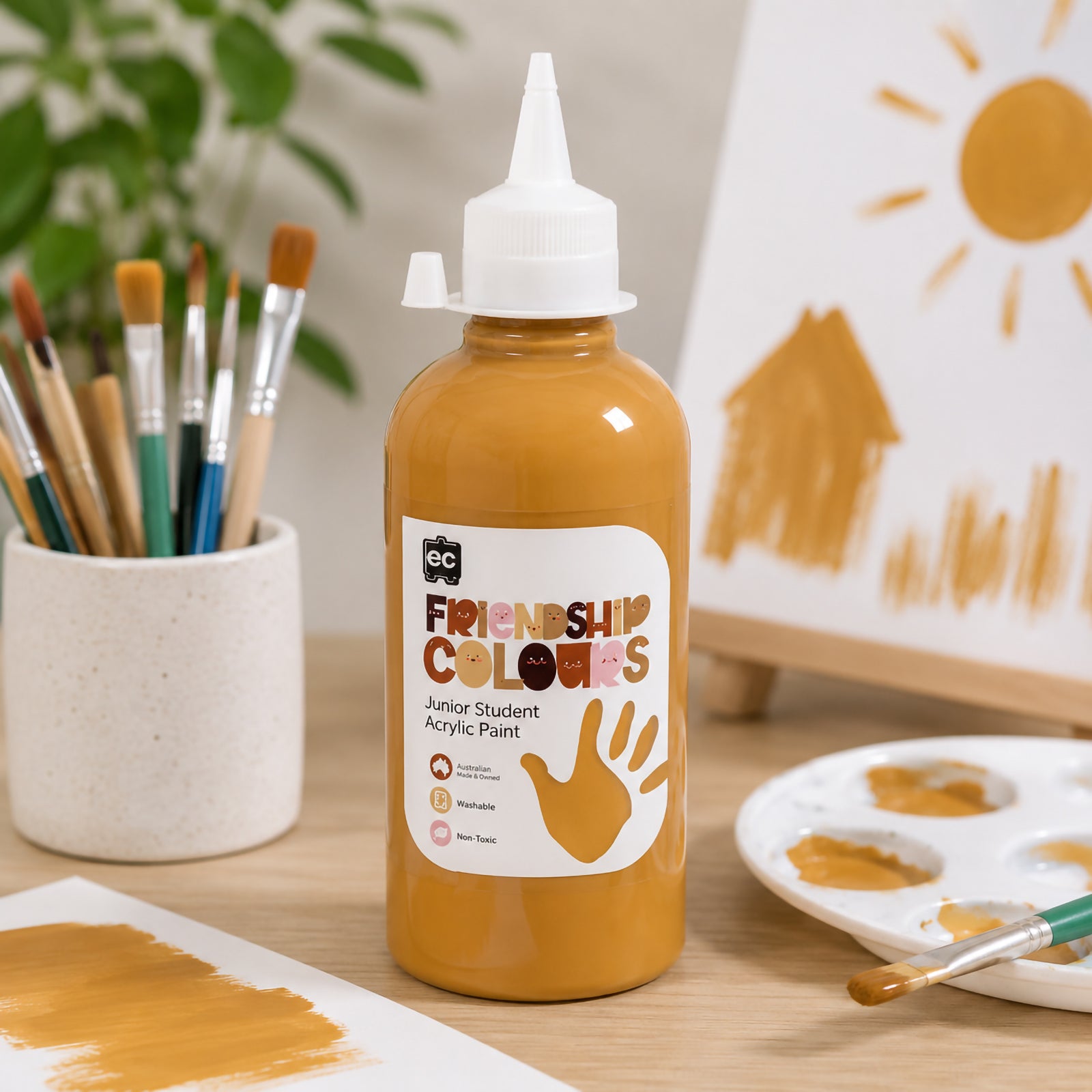 Friendship Colours Honey Acrylic Paint bottle positioned on an art table with brushes, paint palette and a simple painted house scene in the background. The rich golden honey shade demonstrates smooth application and is ideal for classroom creativity and self expression.
