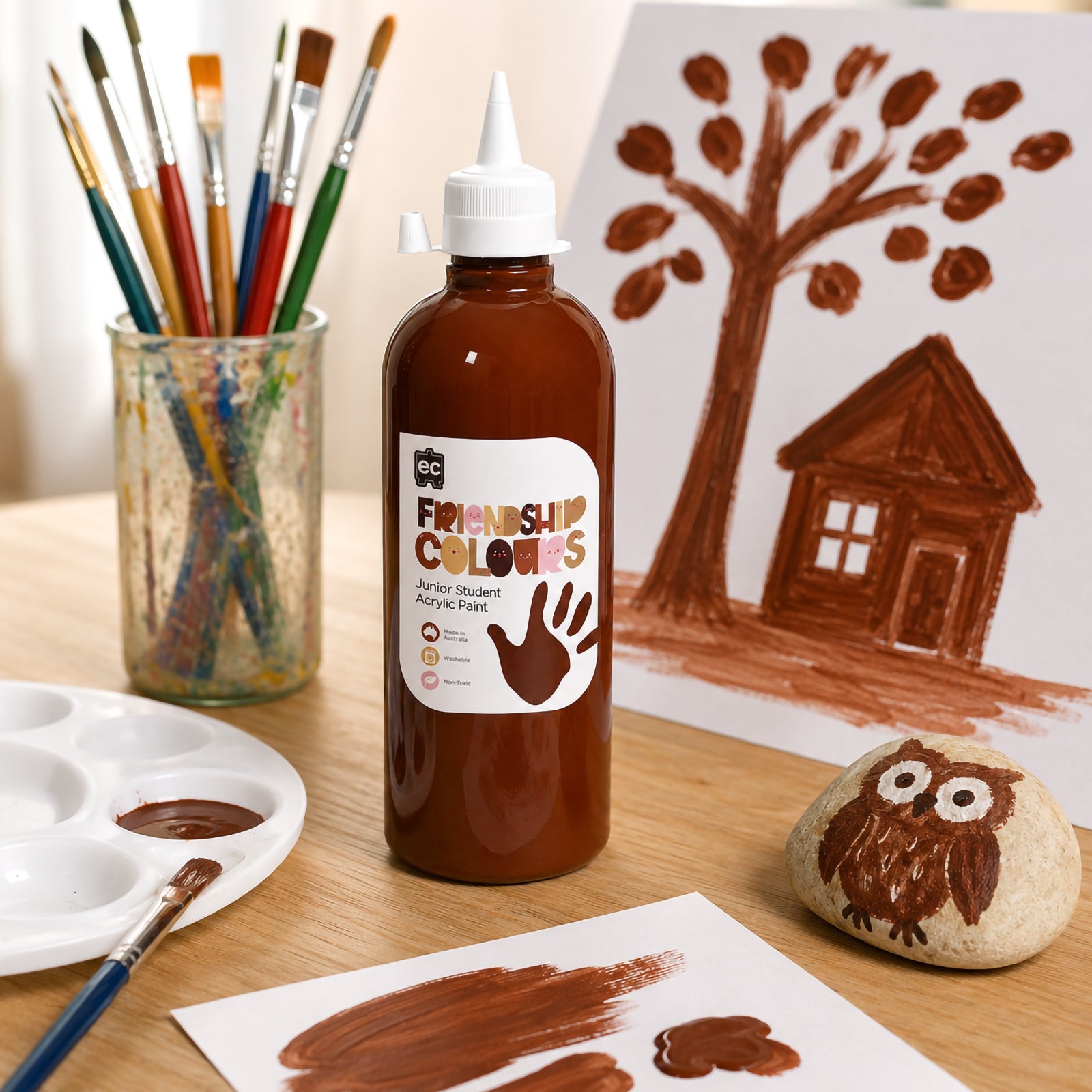 Bottle of Friendship Colours Mahogany Acrylic Paint surrounded by painting tools, artwork and a hand painted decorative stone. The scene showcases the paint's versatility for classroom crafts, mixed media projects and creative learning experiences.