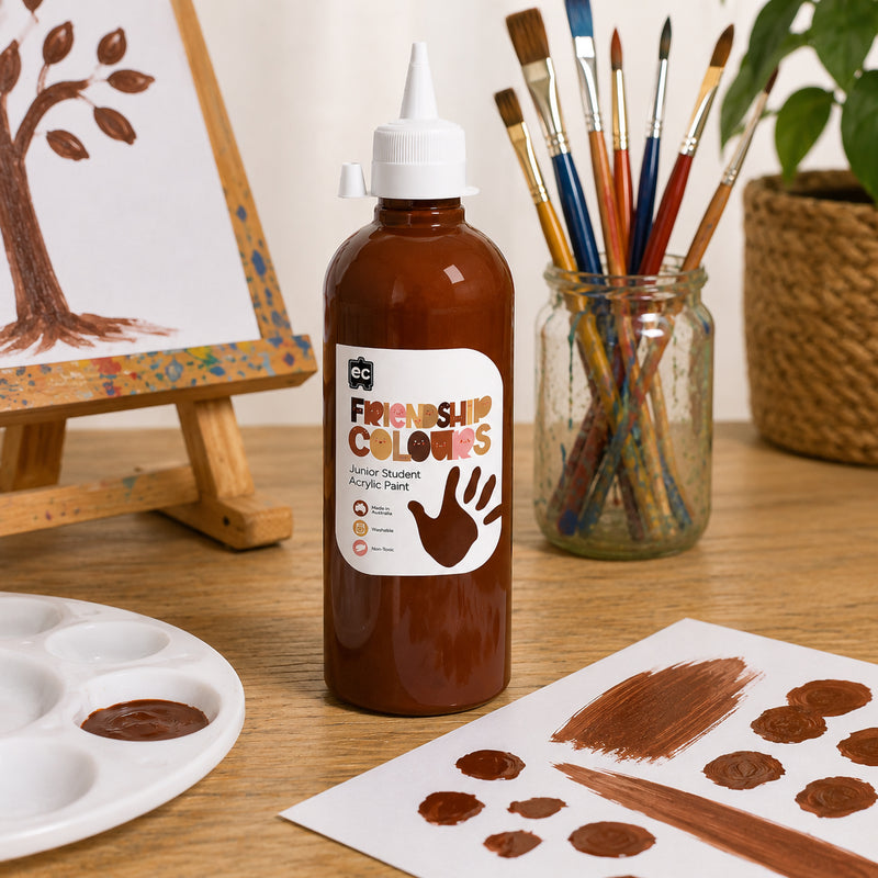 Mahogany Friendship Colours Paint 500ml positioned next to colourful paintbrushes and an easel featuring a tree artwork. The deep skin tone inspired paint demonstrates smooth coverage and is presented as an ideal resource for educational art programs and self expression activities.