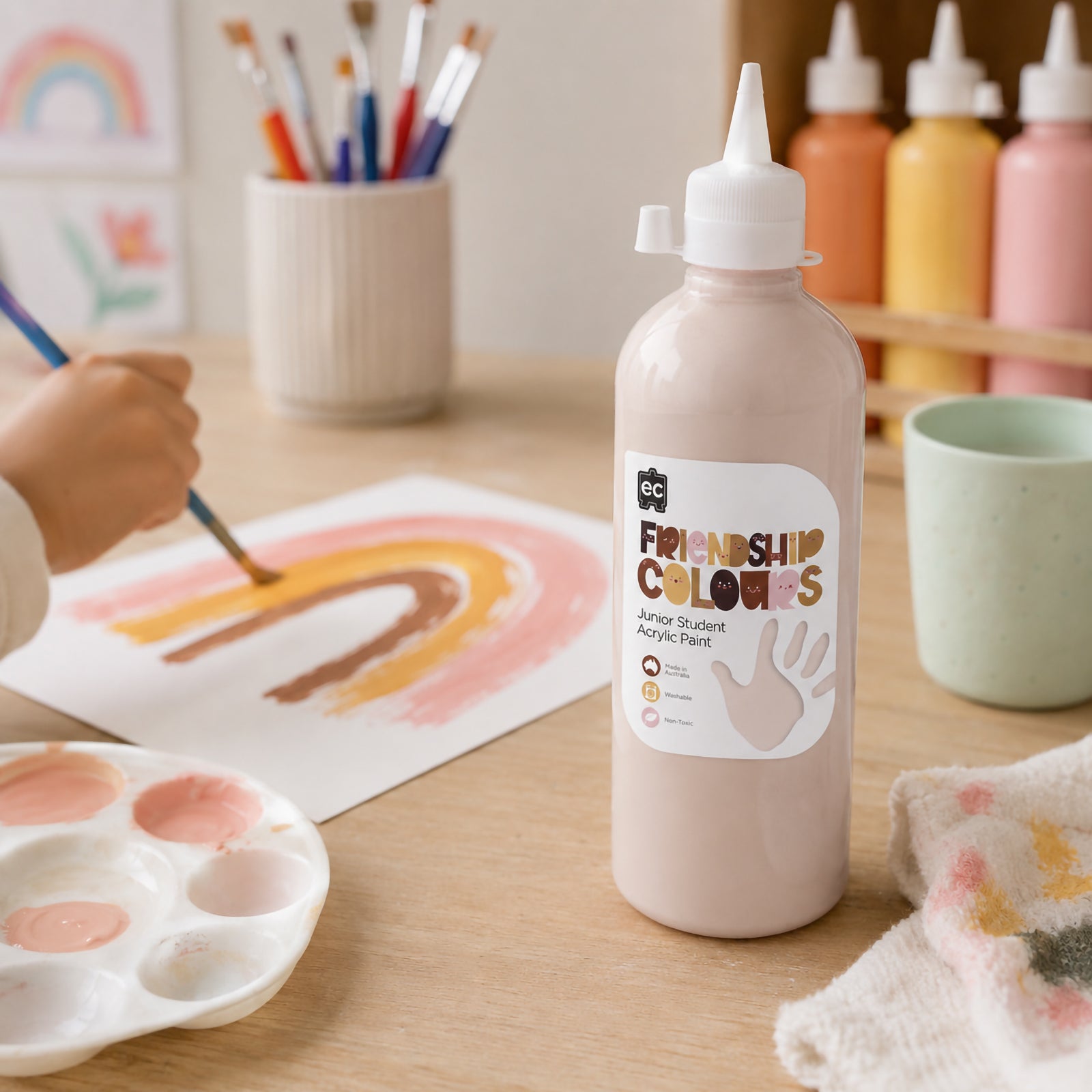 Bottle of Peach Acrylic Paint featured beside a painted rainbow artwork, brushes and classroom paint supplies. The scene highlights creative learning, colour exploration and the use of inclusive skin tone paints for portraits, storytelling and imaginative artwork.