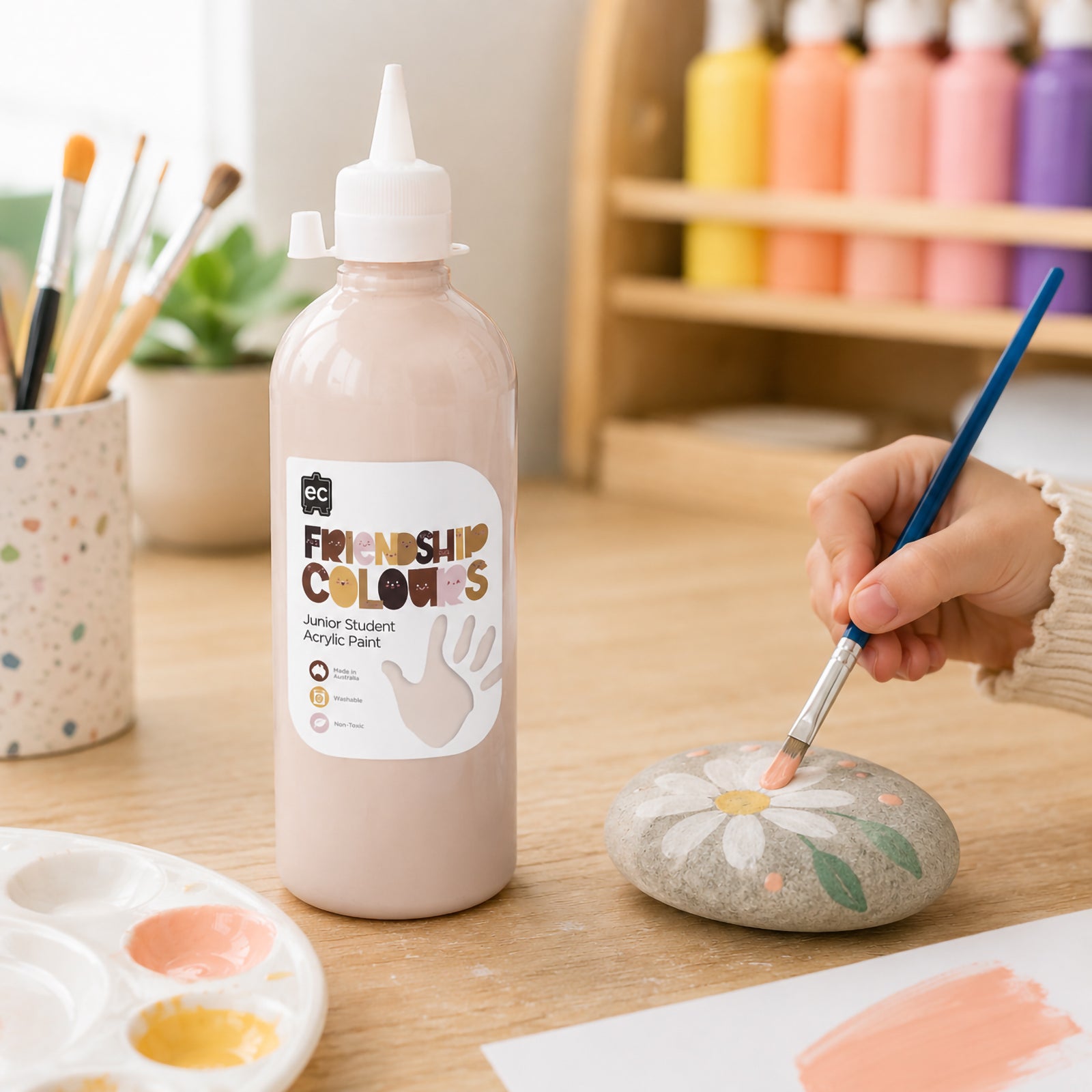 Friendship Colours Peach Paint 500ml positioned on a classroom table while a child paints a decorative flower design on a smooth stone. The image demonstrates the paint's creamy consistency, gentle skin tone shade and suitability for detailed craft and educational art projects.