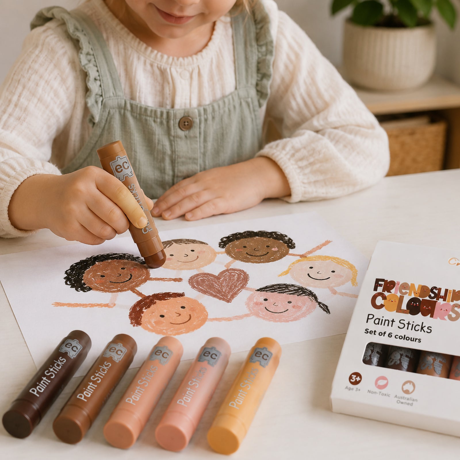 Child using Friendship Colours Paint Sticks to draw a group of smiling faces connected around a heart. Six skin tone inspired paint sticks are displayed alongside the artwork, highlighting inclusive creativity, self expression and multicultural learning activities in an early childhood setting.