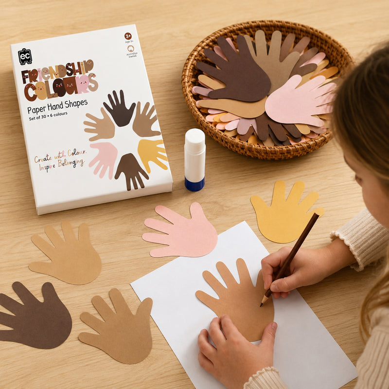 Friendship Colours Paper Hand Shapes packaging displayed beside a classroom craft activity. A child decorates one of the large paper hand cut outs while additional shapes in assorted skin tones are arranged on the table, highlighting opportunities for creative and inclusive learning.