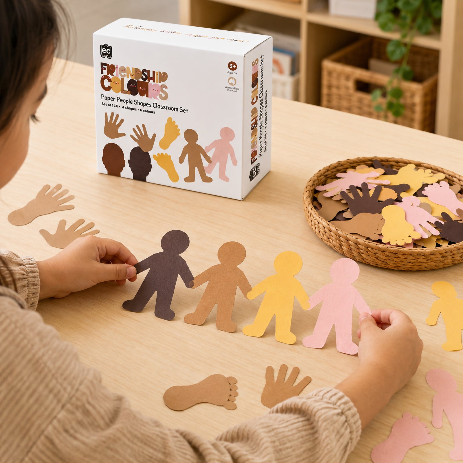 Student engaging in a classroom craft activity with Friendship Colours Paper People Shapes. The educational resource box is displayed alongside assorted paper shapes in realistic skin tones, supporting social awareness, creativity and diversity focused learning projects.