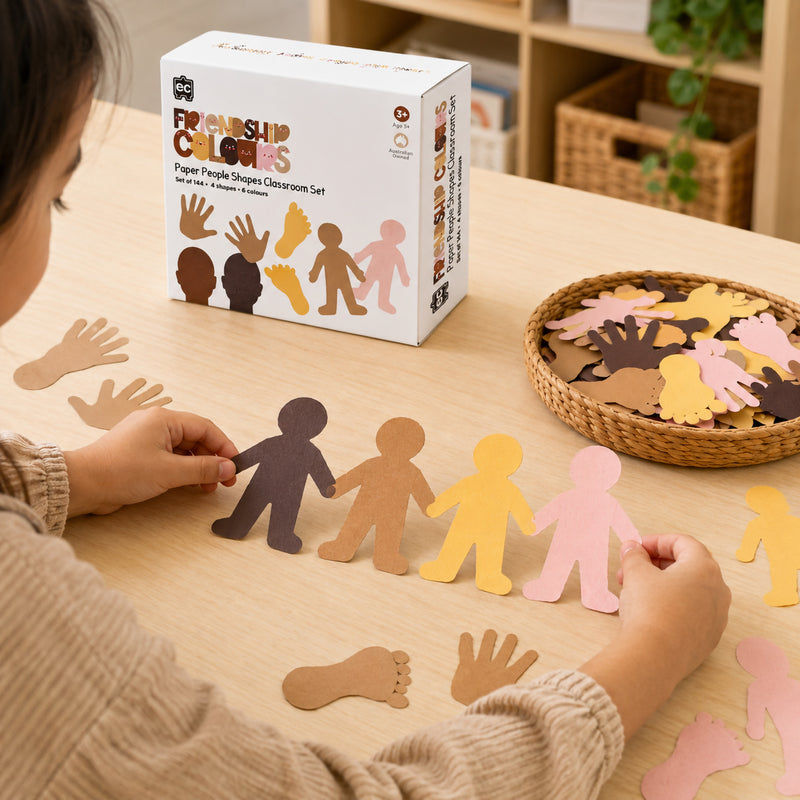Student engaging in a classroom craft activity with Friendship Colours Paper People Shapes. The educational resource box is displayed alongside assorted paper shapes in realistic skin tones, supporting social awareness, creativity and diversity focused learning projects.